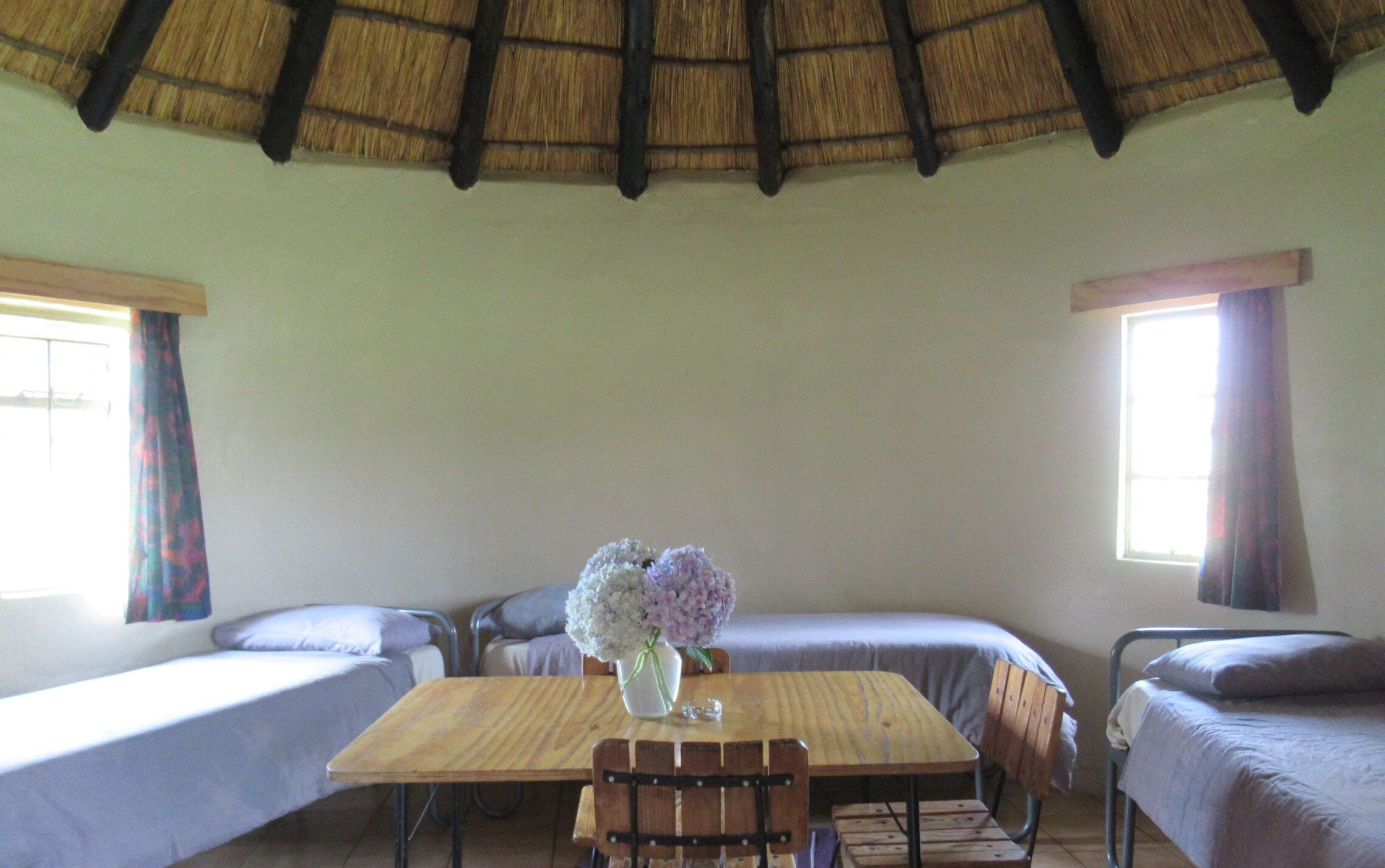 Una habitación con una mesa de madera, dos camas, un jarrón con flores y un techo de paja.