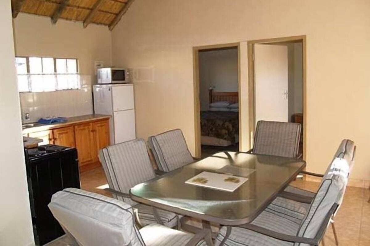 A compact room with a glass table and chairs, a kitchenette with a refrigerator and microwave, and a doorway leading to a bedroom.