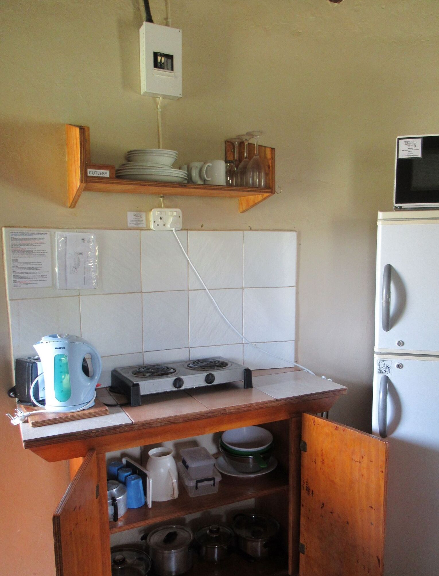 A compact kitchen with a stove, kettle, and a microwave on a small countertop. There is a wooden shelf above the stove holding dishes and glasses. A wooden cabinet below the shelf contains pots and a kettle.