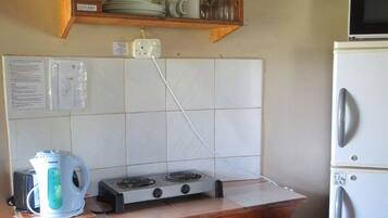 A compact kitchen with a stove, kettle, and a microwave on a small countertop. There is a wooden shelf above the stove holding dishes and glasses. A wooden cabinet below the shelf contains pots and a kettle.