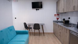 Ett kompakt vardagsrum med en turkosfärgad soffa, ett matbord med två stolar, ett köksområde med träskåp och en inbyggd ugn samt en TV som är monterad på väggen.
