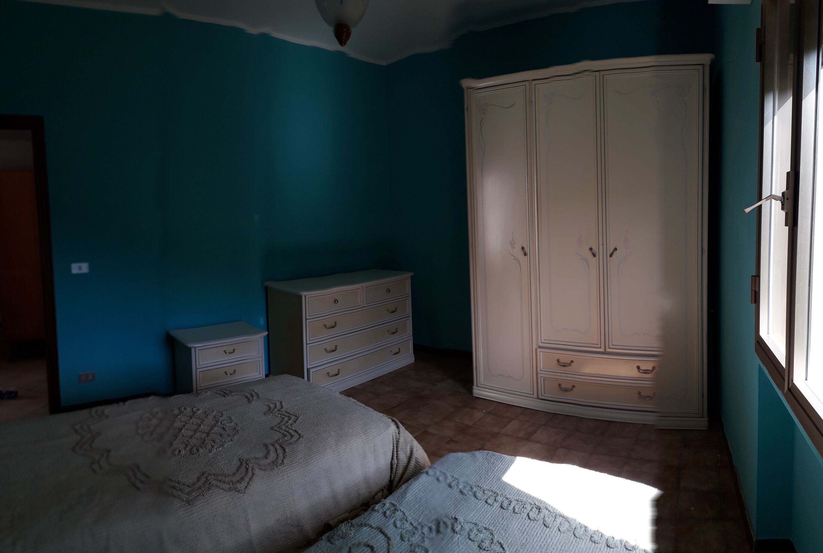 A bedroom with teal walls, a large wardrobe, a bed with patterned bedding, and a small dresser.