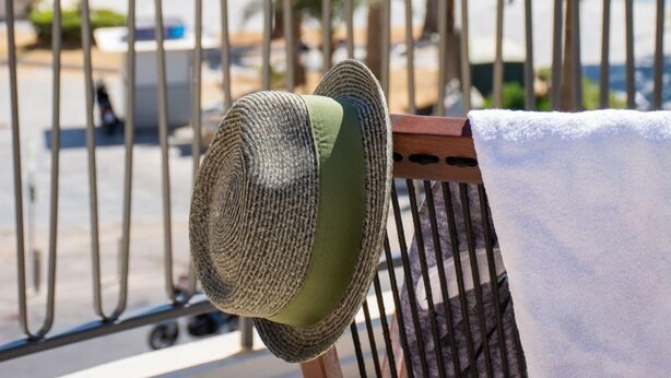 A balcony with a hat and towel, overlooking a coastal area with a lighthouse and palm trees.