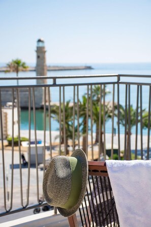 A balcony with a hat and towel, overlooking a coastal area with a lighthouse and palm trees. - V77 Seafront Leto Suite with roof top hot tub. (Rethymno)