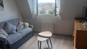 A cozy loft bedroom with a dark blue sofa, a small round table, and a view of buildings through a window. - Bright, functional apartment by the sea (Villers-sur-Mer)