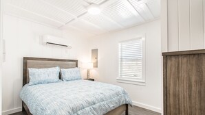 Ein Schlafzimmer mit einem Bett, einer Klimaanlage, einem Nachttisch mit Lampe, einem Fenster mit Jalousien und einem hölzernen Kopfteil.