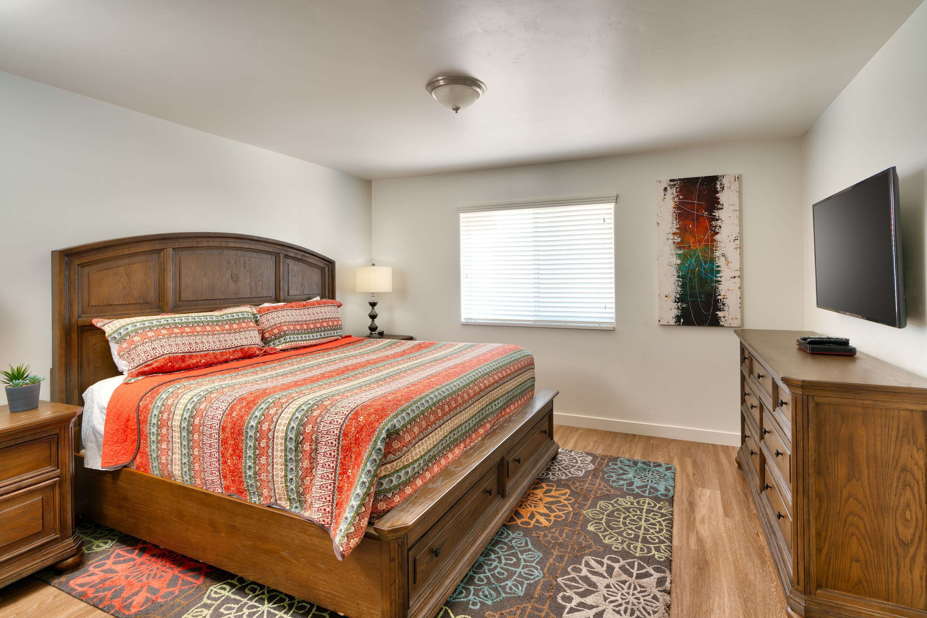 Une chambre à coucher avec un lit en bois, une table de chevet, une commode, un téléviseur et une fenêtre avec des stores.