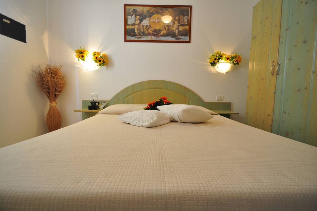 A neatly made bed with a quilted cover, a vase with dried plants, and wall-mounted lamps with sunflowers.