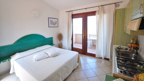 A bedroom with a bed, a window with curtains, a view of the outside, and a kitchenette with a stove. - Residenze La Corte (Badesi)
