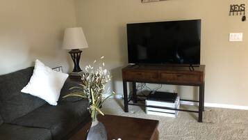 A living room with a dark-colored sofa, a wooden coffee table with a vase of flowers, a flat-screen TV on a wooden stand, and a lamp on a side table.