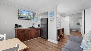 A modern kitchen with wooden cabinets, a stainless steel refrigerator, and a flat-screen TV mounted above the counter. - Pelican Pointe 429:Walk to Beach~Pool~Free Parking (Clearwater Beach)