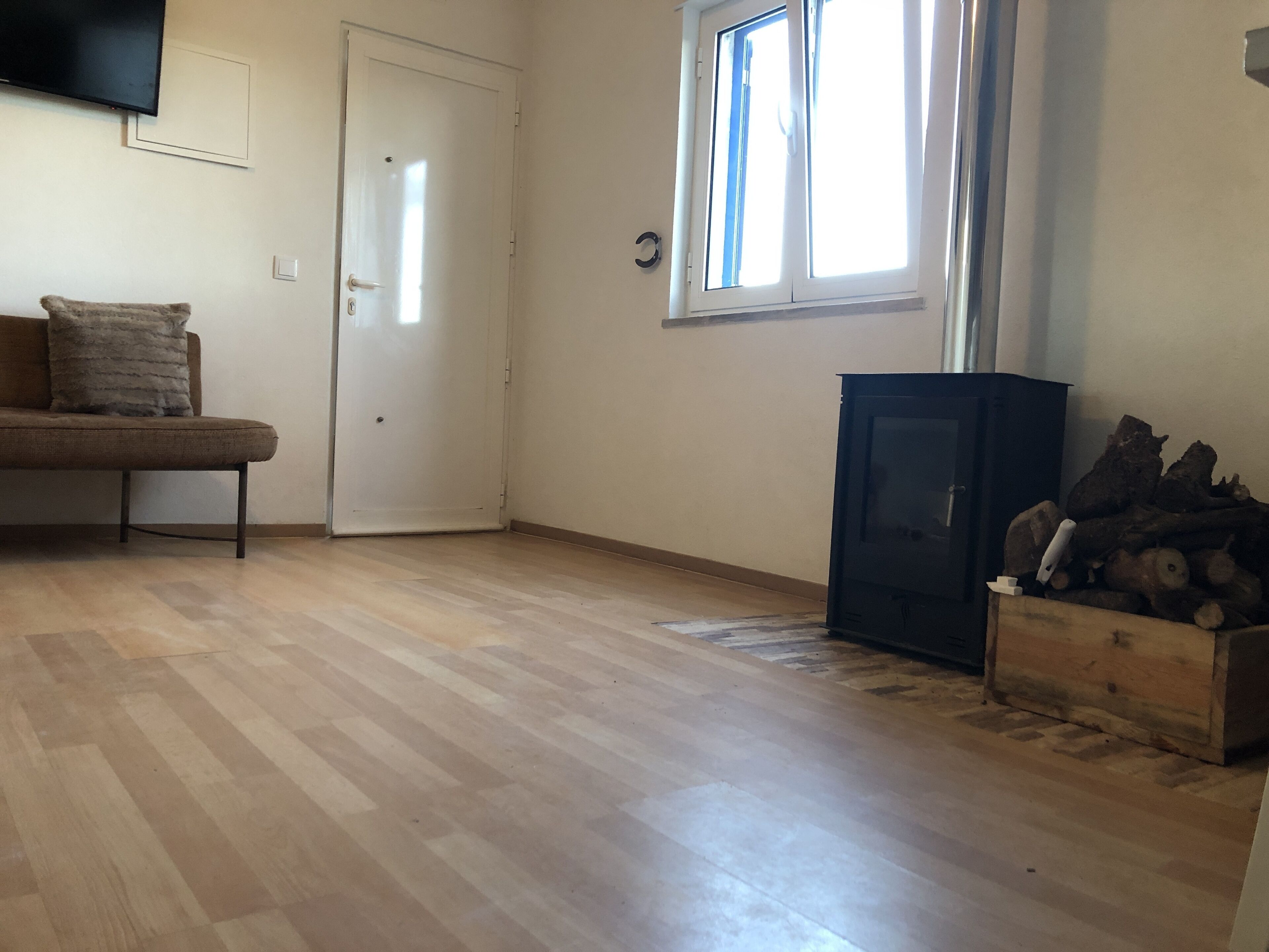 Een kamer met houten vloer, een bank met kussen, een aan de muur bevestigde televisie, een raam met zonwering en een houten kast met houtblokken.