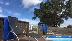 Deux chaises longues placées près d’une piscine, avec une terrasse en bois et un grand arbre en arrière-plan.
