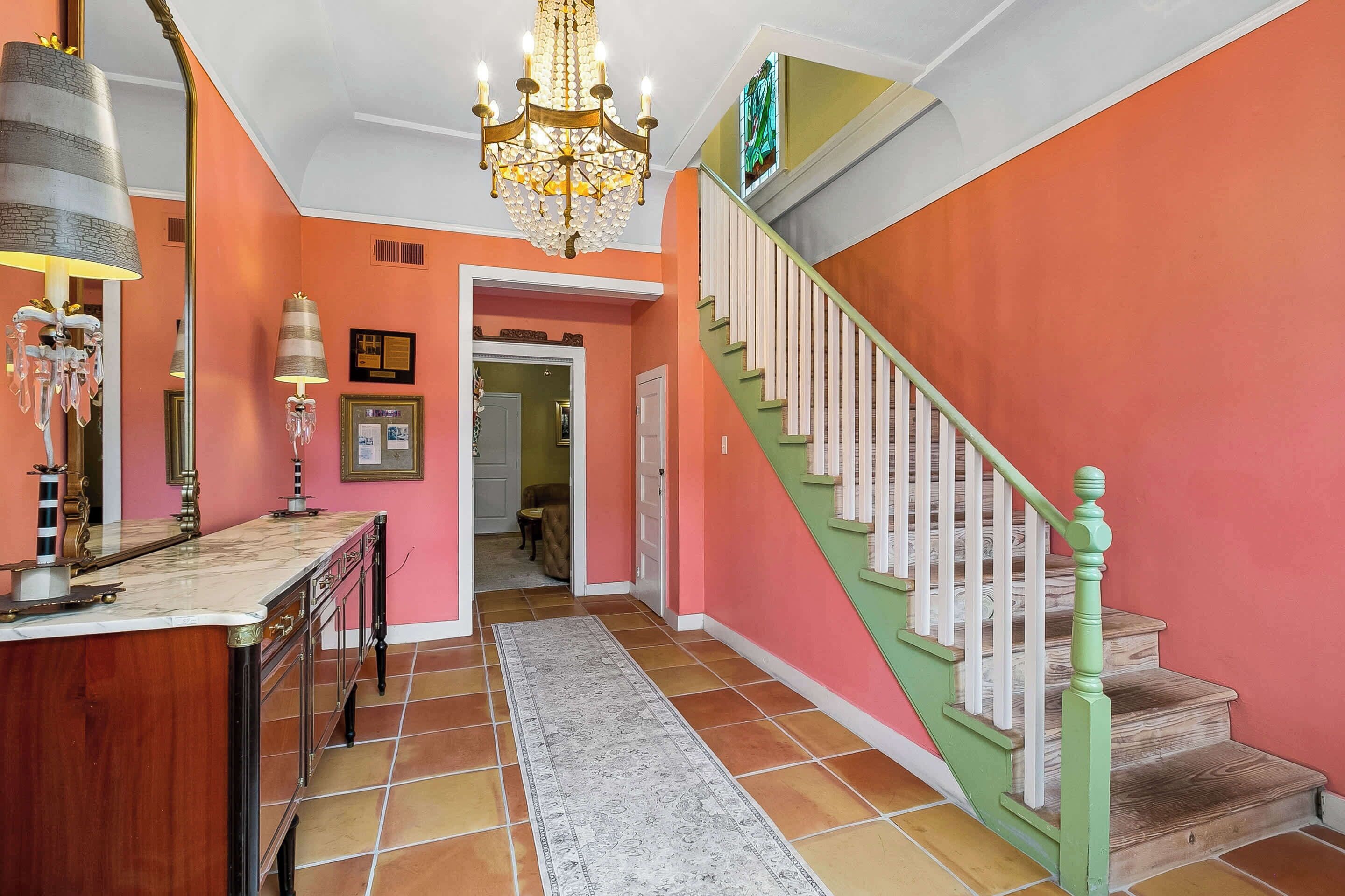 Un couloir avec un comptoir en marbre, un lustre, un escalier avec des rampes vertes et un sol carrelé.