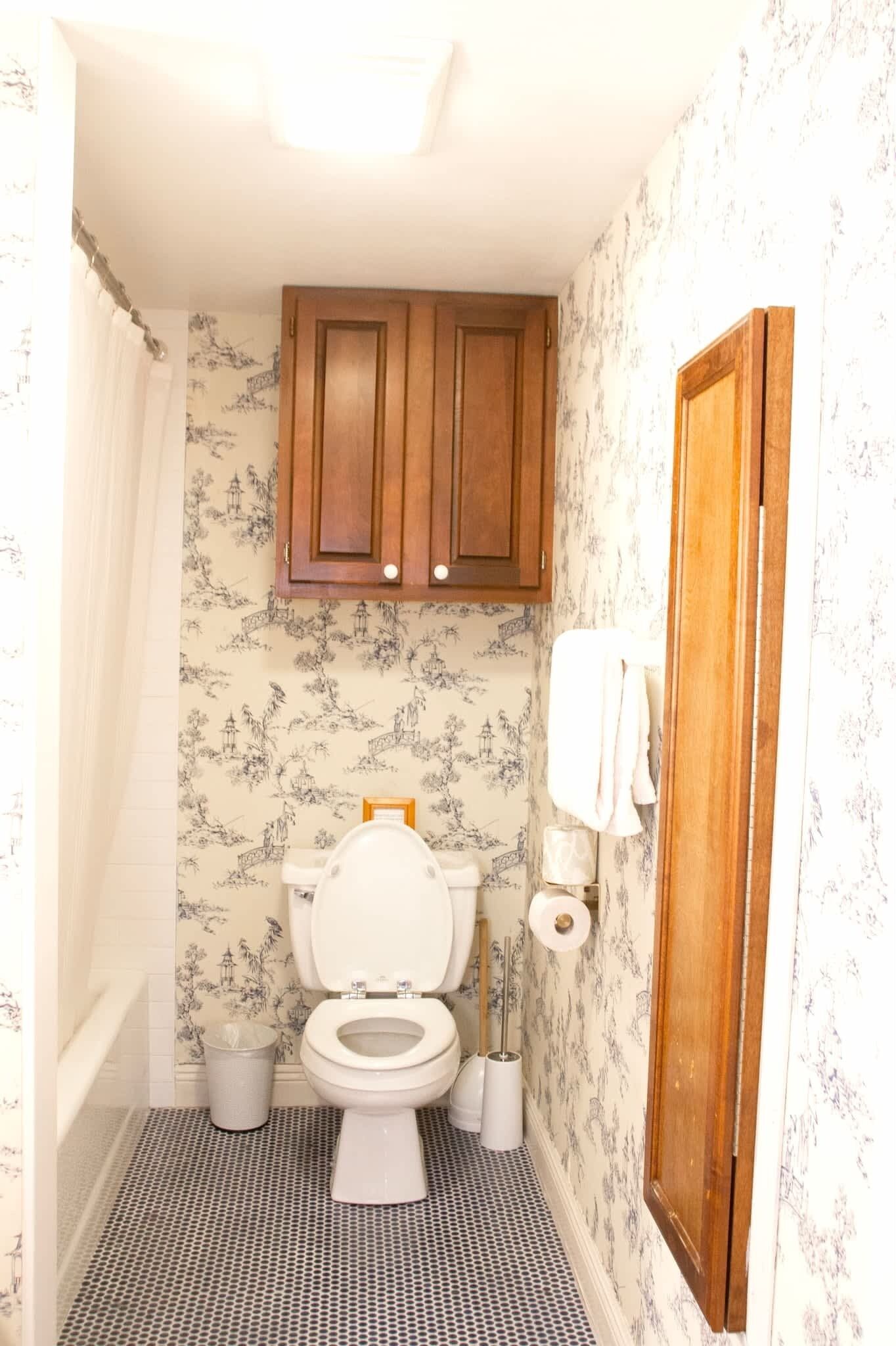 Een badkamer met een toilet, houten kastjes, behang met een patroon en een douchegordijn.