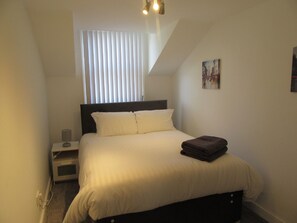 Un dormitorio con cama, mesita de noche, lámpara, ventana con persianas y un cuadro en la pared.