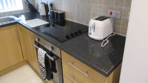 Una cocina con encimera negra, un tostador, una tetera y una cafetera. Hay armarios de madera y un salpicadero de azulejos.