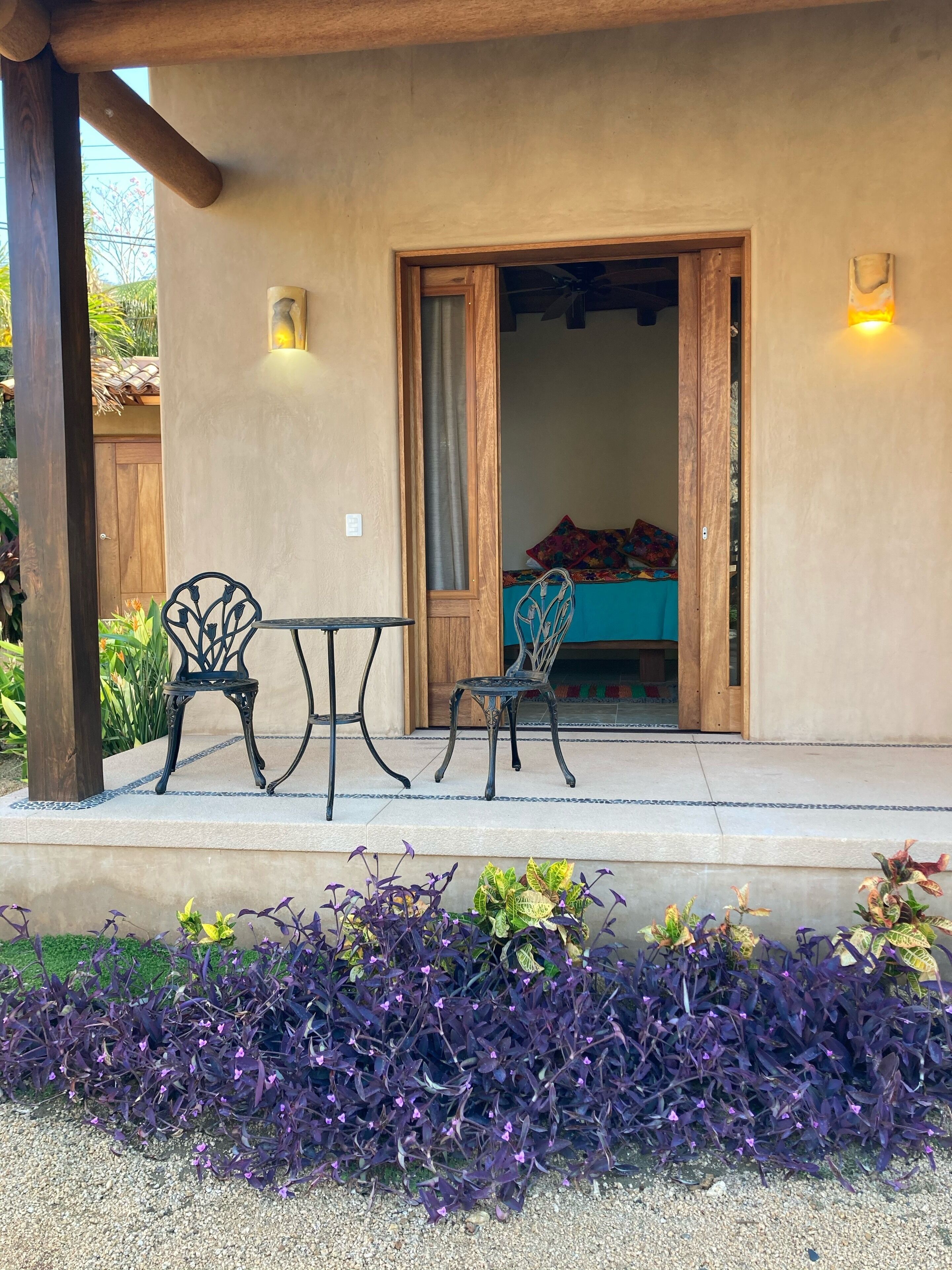 Un porche cubierto con dos sillas y una mesa, una puerta de madera que da al interior, y un pequeño jardín con follaje morado.