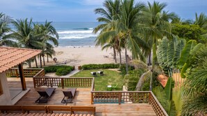 Blick auf einen Strand von einer Holzterrasse mit Liegestühlen, einem kleinen Tisch und Ausblick auf das Meer und Palmen.