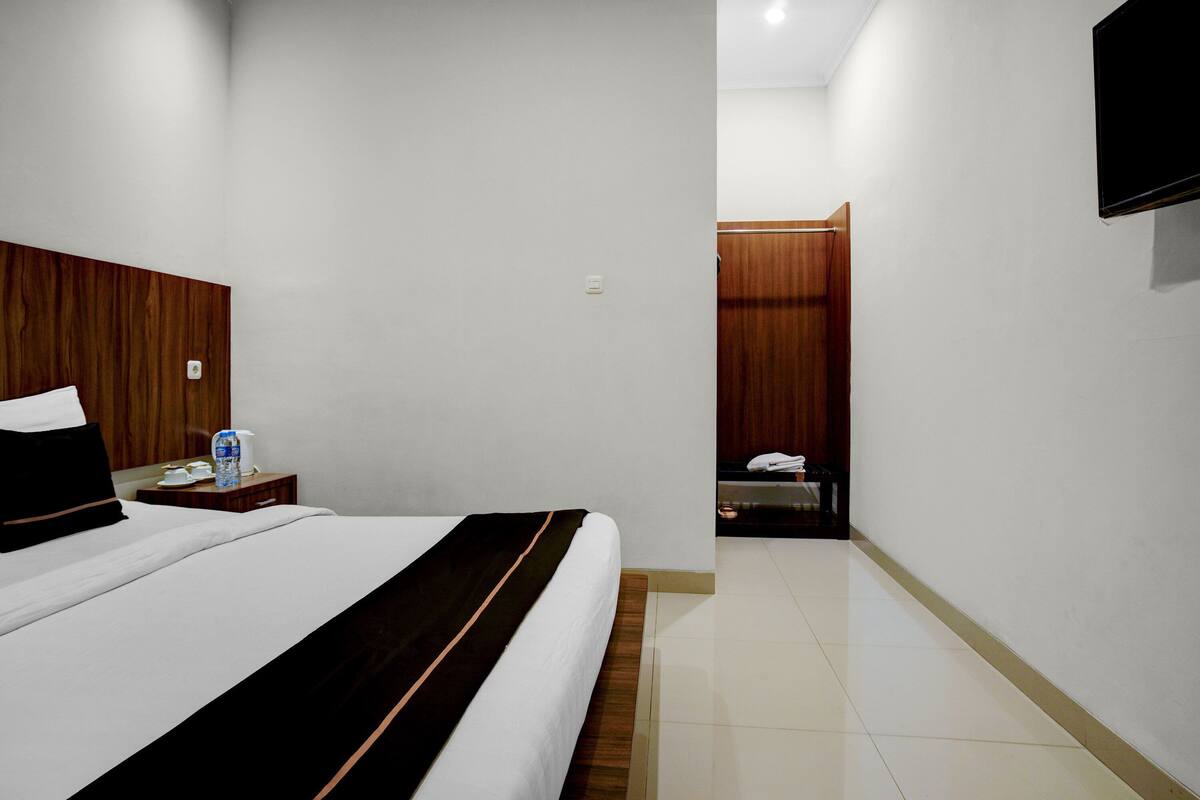 Une chambre d’hôtel avec un lit, une tête de lit en bois, un téléviseur fixé au mur et une salle de bain visible à travers une porte ouverte.