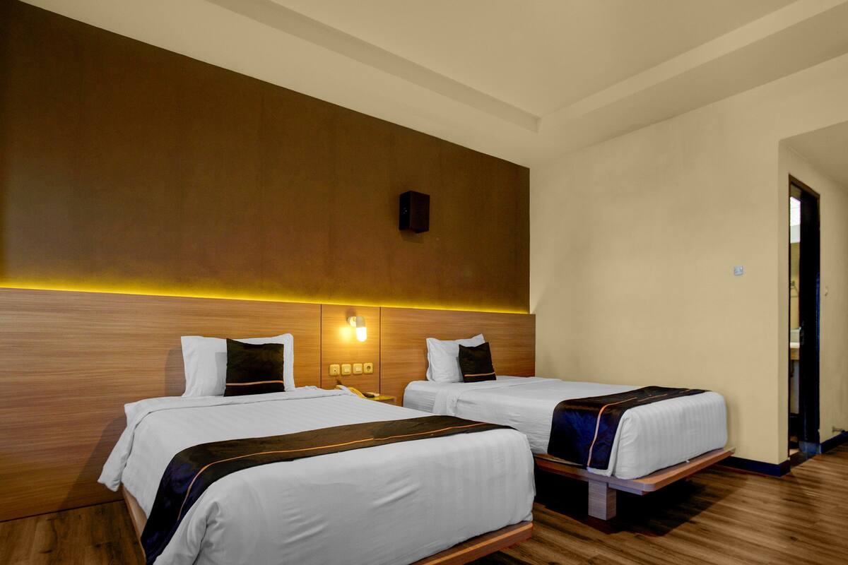 Une chambre d’hôtel avec deux lits, une tête de lit en bois, une lampe fixée au mur et une porte.