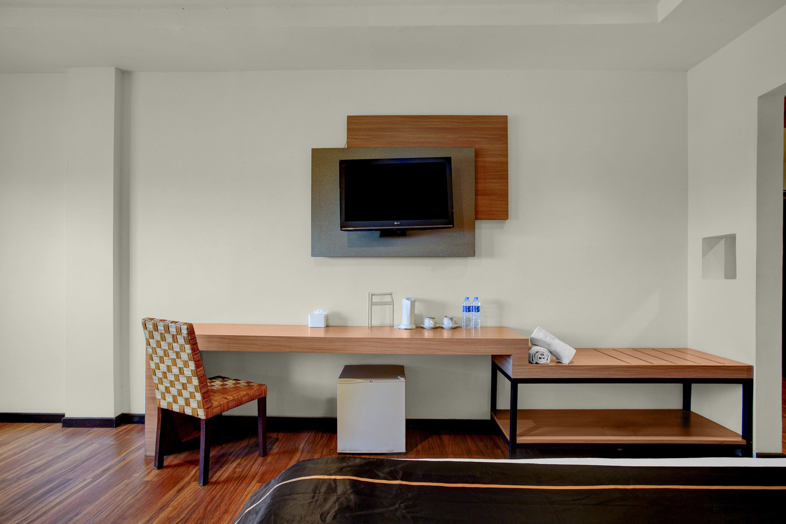 Quarto de hotel com uma escrivaninha de madeira, televisão fixada na parede, uma cadeira, um pequeno frigorífico e uma cama.