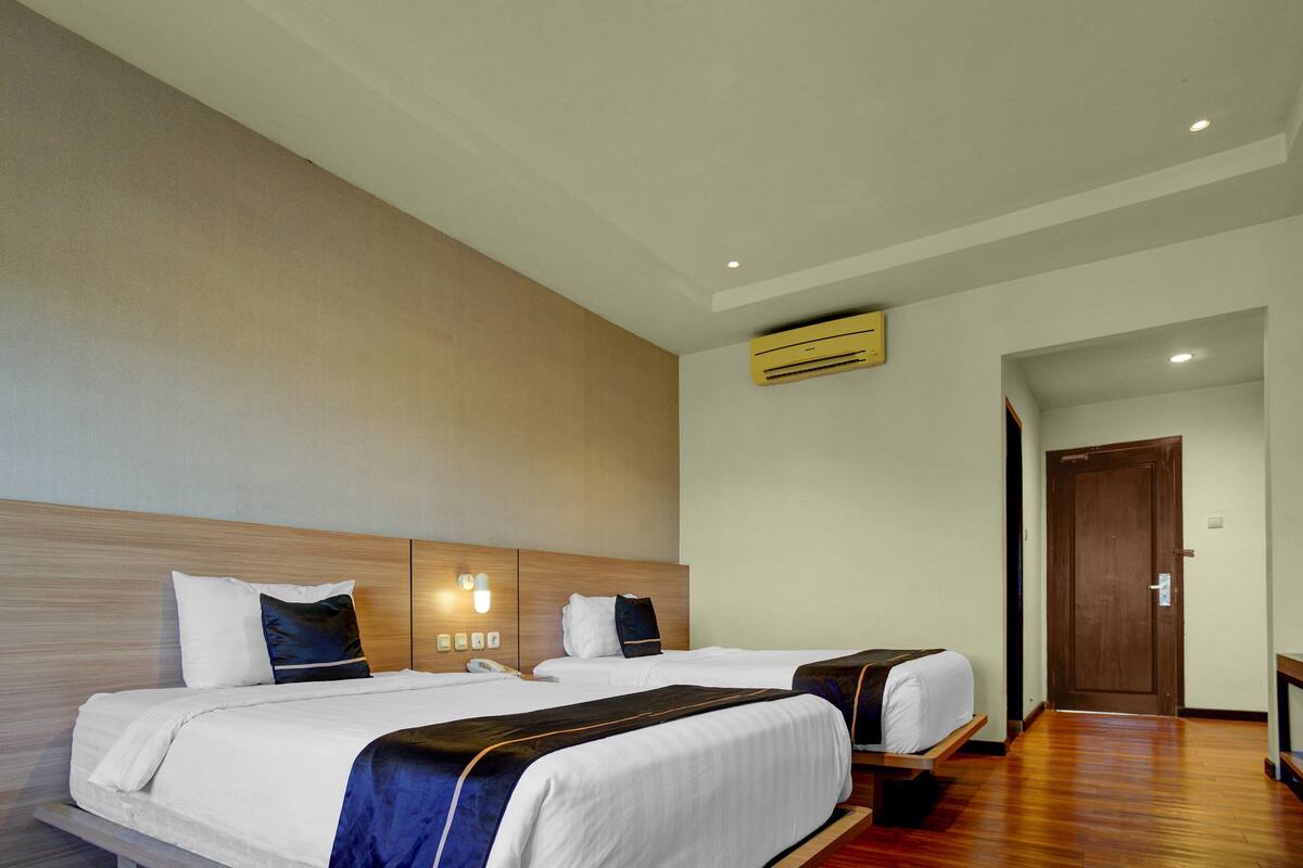 Une chambre d’hôtel avec deux lits, une tête de lit en bois, un climatiseur fixé au mur et une porte donnant sur une autre chambre.