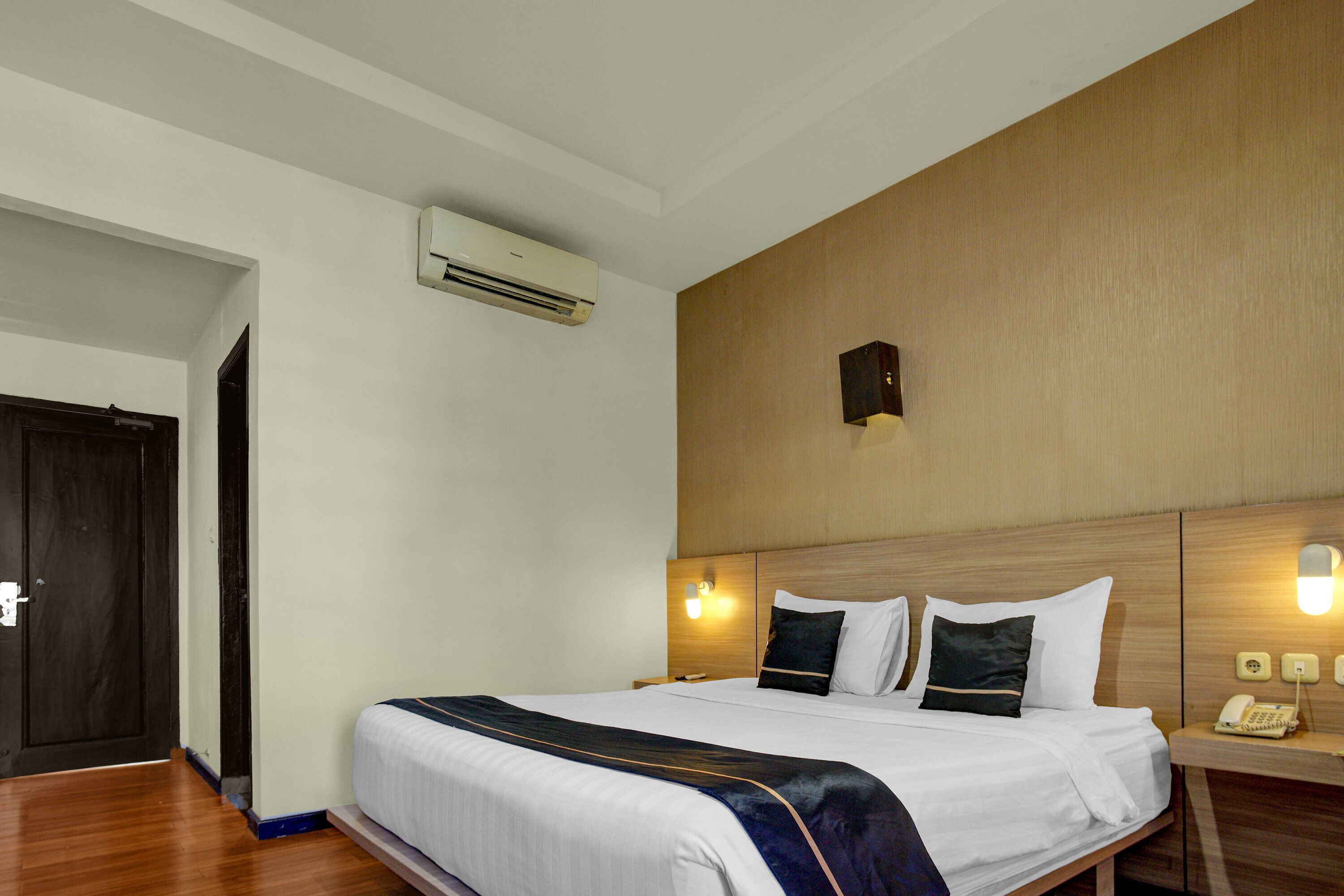 Quarto de hotel com uma cama grande, ar condicionado, mesa de cabeceira com abajur e cabeceira de madeira.