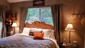 A bedroom with a large bed, a headboard, two bedside lamps, a nightstand, a vase with flowers, and a window with curtains.