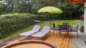 Une terrasse en bois avec des chaises longues et une table, entourée d’une haie et d’arbres.