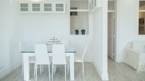 Een moderne eethoek met een glazen tafel en witte stoelen, een plantje op een plank, en een witte bank in de aangrenzende kamer.