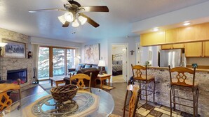 A spacious living area with a fireplace, a dining table with chairs, and a kitchen with wooden cabinets and a refrigerator. - Walk to lifts | Easy Access | Wi-Fi | Mountain Spirits 215 (Angel Fire)