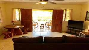 A living room with a sofa, a TV, a dining table with chairs, and a view through open doors to an outdoor area. - Tropical Patio & Garden, Walk to Samara Beach
 (Sámara)