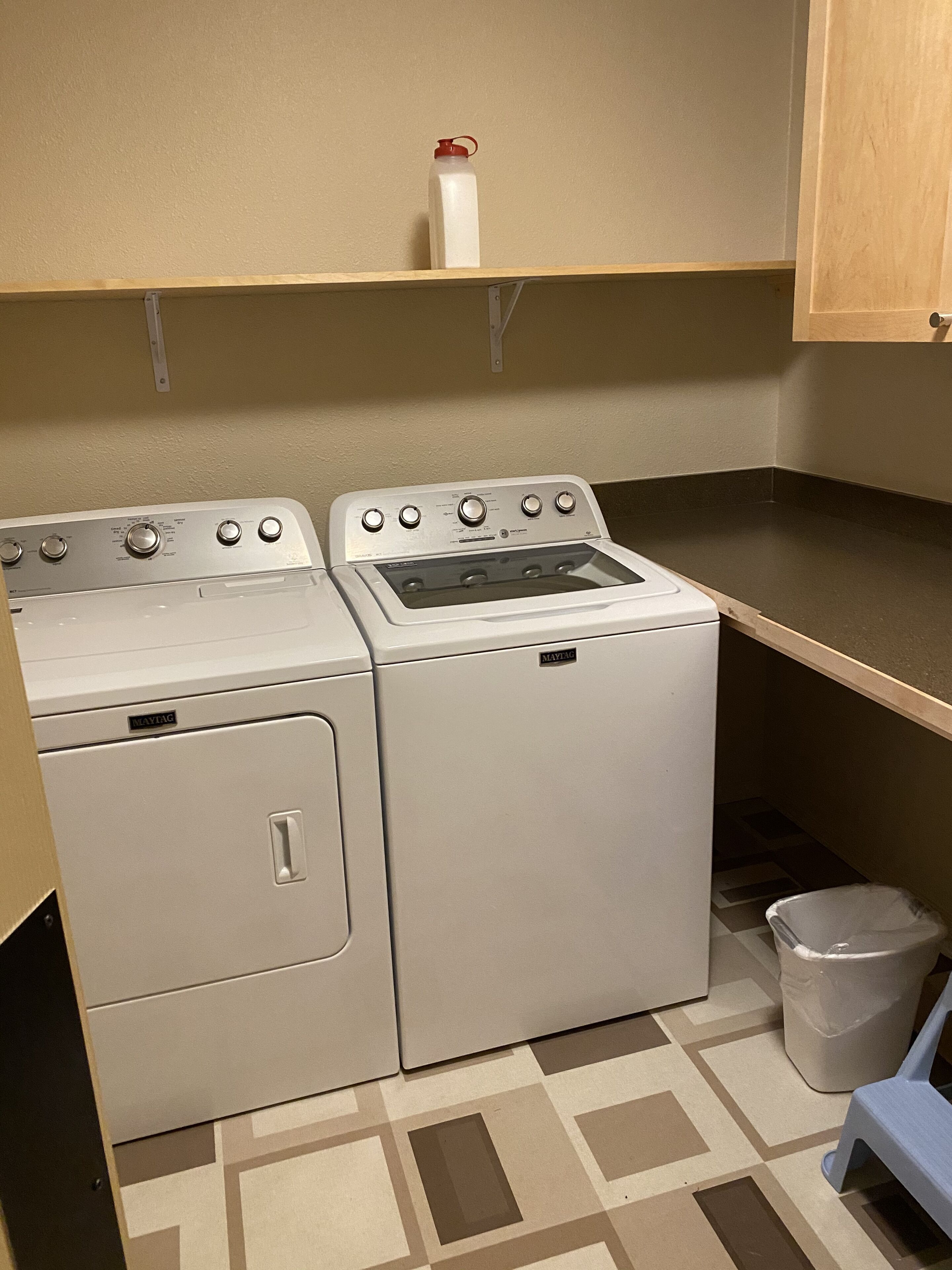 Une buanderie équipée de deux machines à laver et de deux sèche-linge Maytag, d’une petite étagère avec une bouteille d’eau et d’une poubelle.