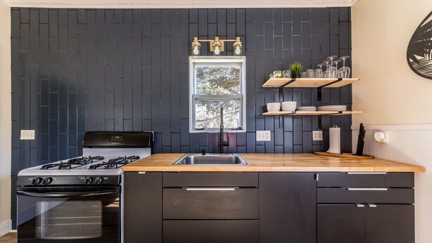 A modern kitchen with a gas stove, oven, and wooden countertop. There is a window with a view of trees, and shelves with kitchenware.