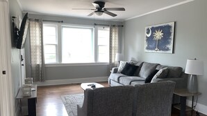 En stue med en grå sofa, et sofabord, et sengebord med en lampe, en loftsvifte og store vinduer med mønstrede gardiner.