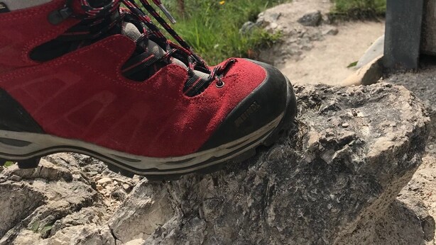 Ein roter Wanderschuh auf felsigem Untergrund, im Hintergrund eine Berglandschaft.