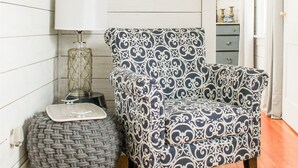 Ein Zimmer mit einem gemusterten Sessel, einem kleinen Hocker, einer Lampe und einem Regal mit Dekoration.