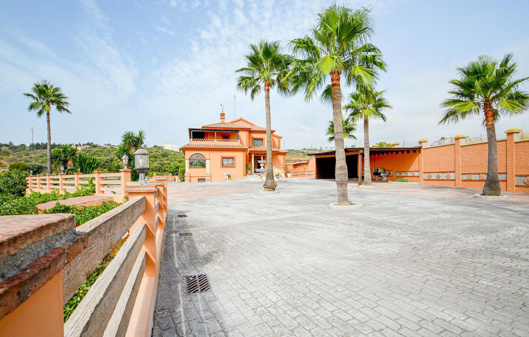 Un amplio patio con un edificio de techo de tejas y una fuente, rodeado de palmeras y un muro de ladrillo.