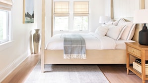 Une chambre à coucher avec un lit, deux fenêtres équipées de stores, une table de chevet avec une lampe et un vase posé sur le sol.