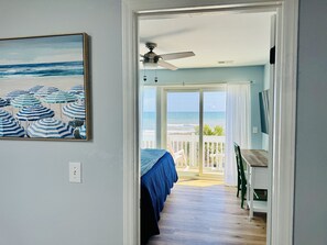 Un dormitori amb llit, ventilador de sostre i vista a la platja per una porta oberta.
