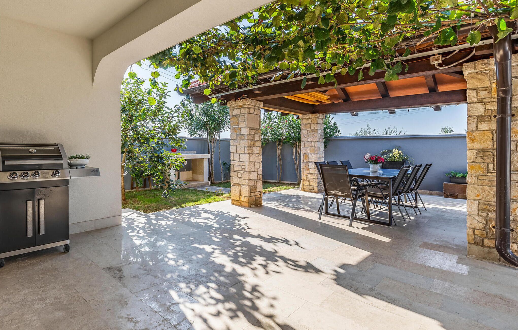Eine Terrasse im Freien mit Grill, Steinpfeilern, einer Pergola mit Kletterpflanzen und einem Tisch mit Stühlen.