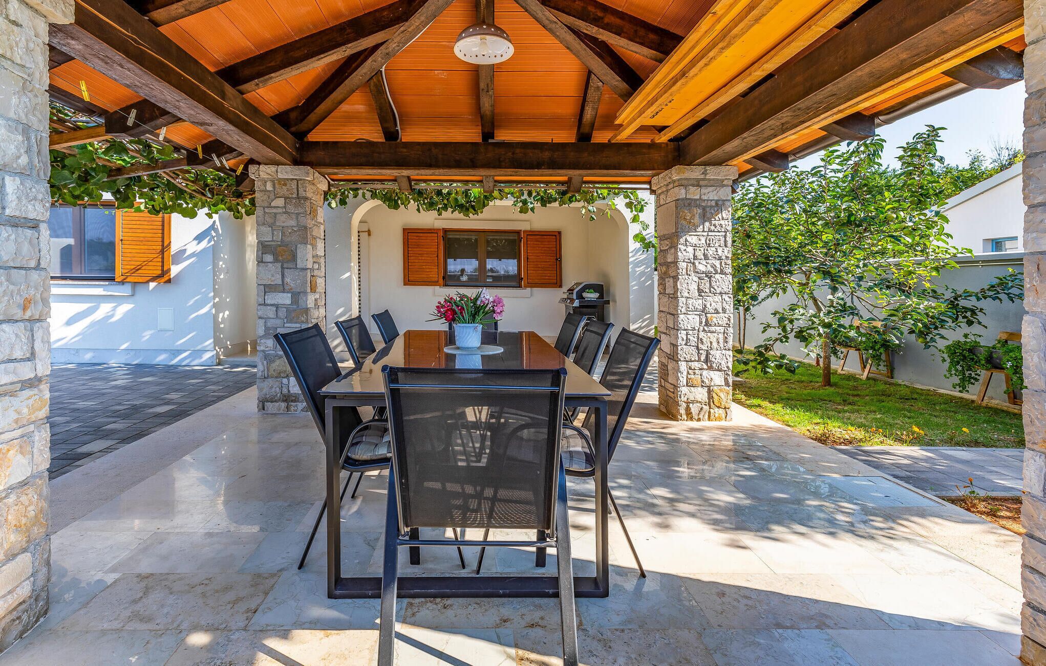 An outdoor covered patio with a stone wall, wooden beams, and a table with chairs.