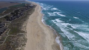 An aerial view of a coastal area with a sandy beach, a road, and a row of houses along the shore.
