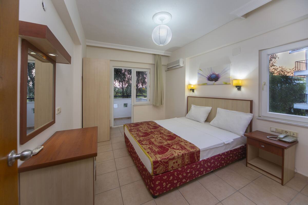A hotel room with a bed, a bedside table with a phone, a window with a view of trees, and a door leading to another room.