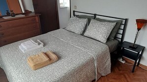 A neatly made bed with patterned bedding, a wooden dresser, a mirror, and a bedside table with a lamp. - Vivenda Unifamiliar (Charneca de Caparica)