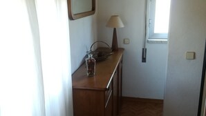 A small, narrow room with a wooden ceiling and a herringbone-patterned floor. There is a wooden counter with a lamp and a vase of flowers on it, and a window with a view of the outside. - Vivenda Unifamiliar (Charneca de Caparica)
