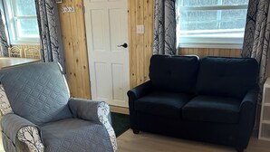 A room with a wooden paneled wall, a white door, a black sofa, and a grey armchair with patterned cushions.