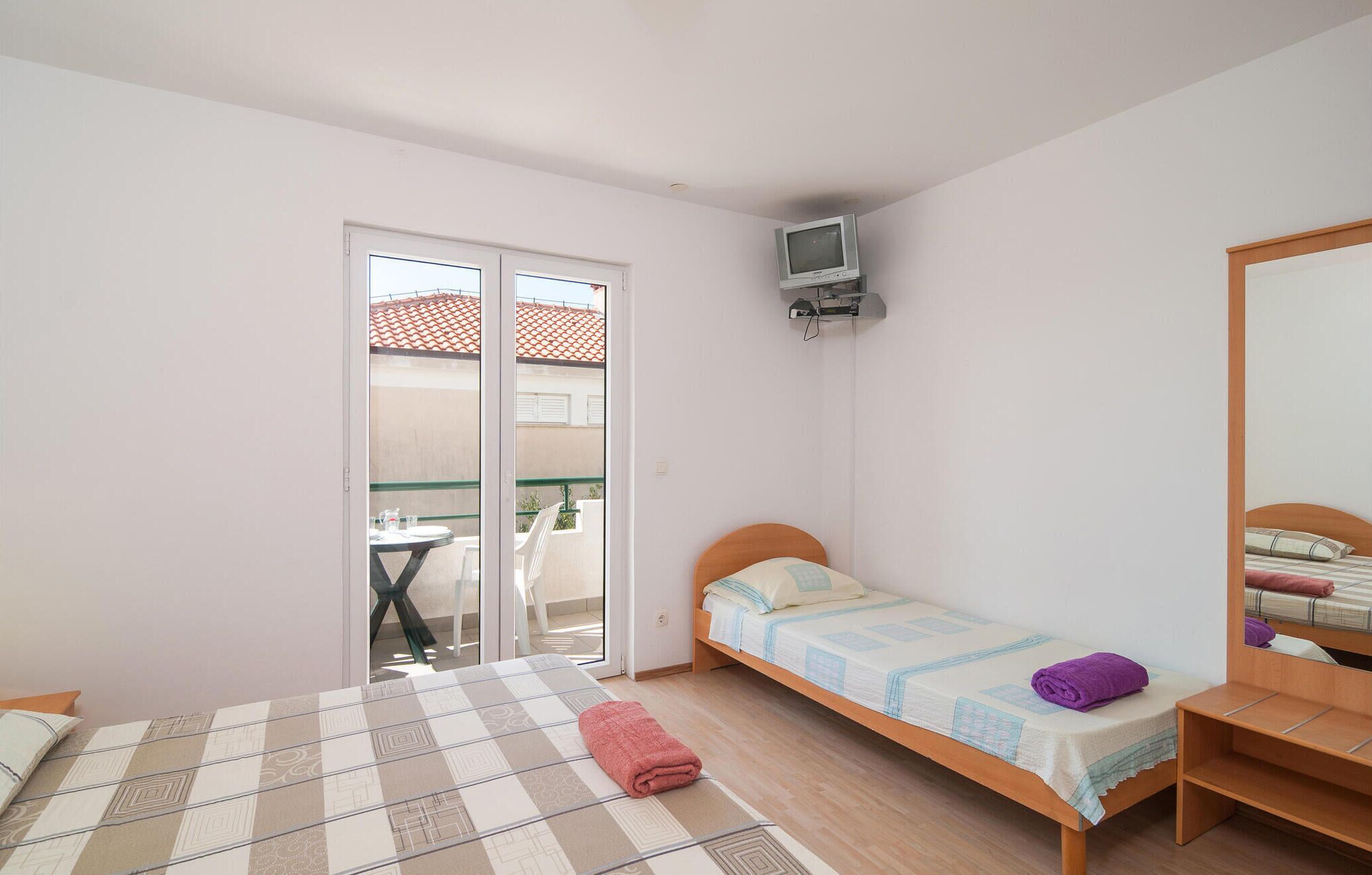 A small, well-lit bedroom with a single bed, a wooden nightstand, a TV mounted on the wall, and a sliding glass door leading to an outdoor area.
