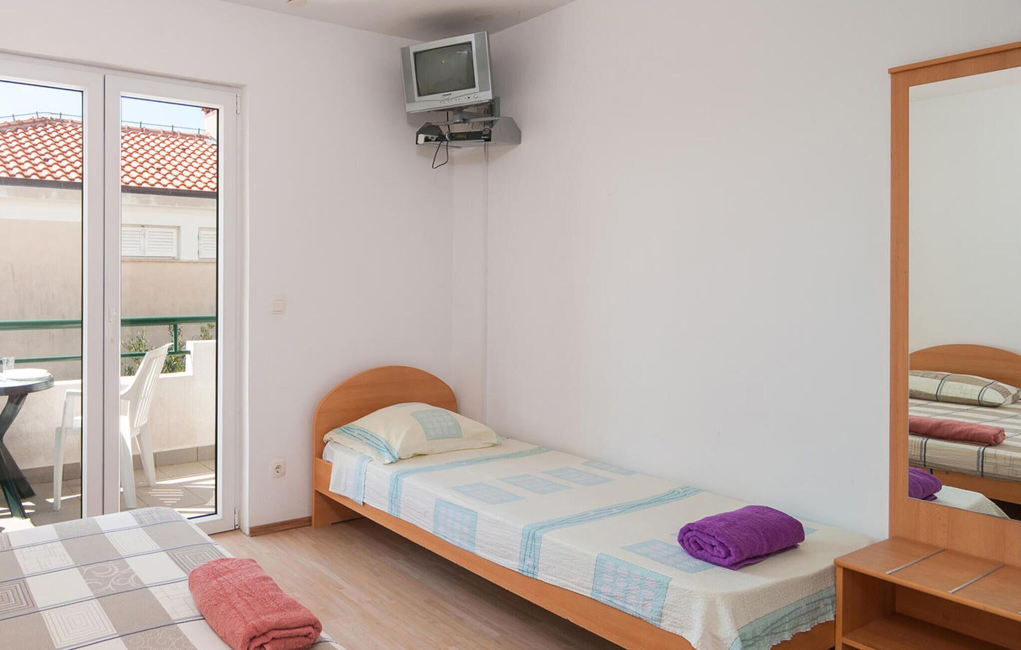 A small, single-bed room with a television mounted on the wall, a balcony with a table and chairs, and a wooden wardrobe.
